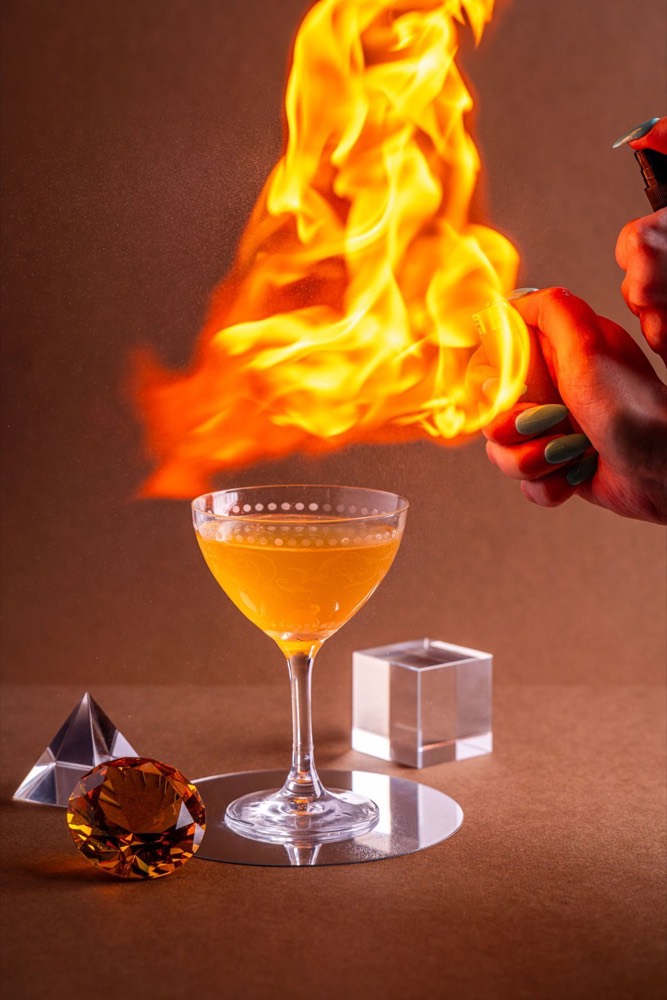 A coupe cocktail being finished with a dramatic flamed orange garnish.