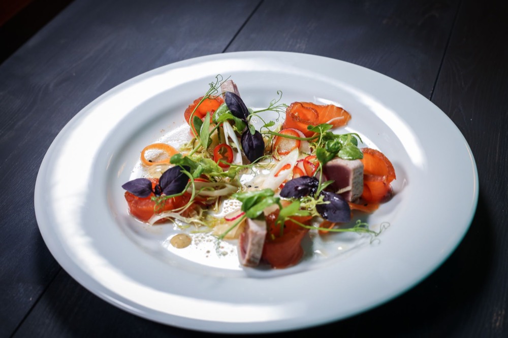 A plated fine-dining dish of cured fish, tomato and micro herbs on a white plate.