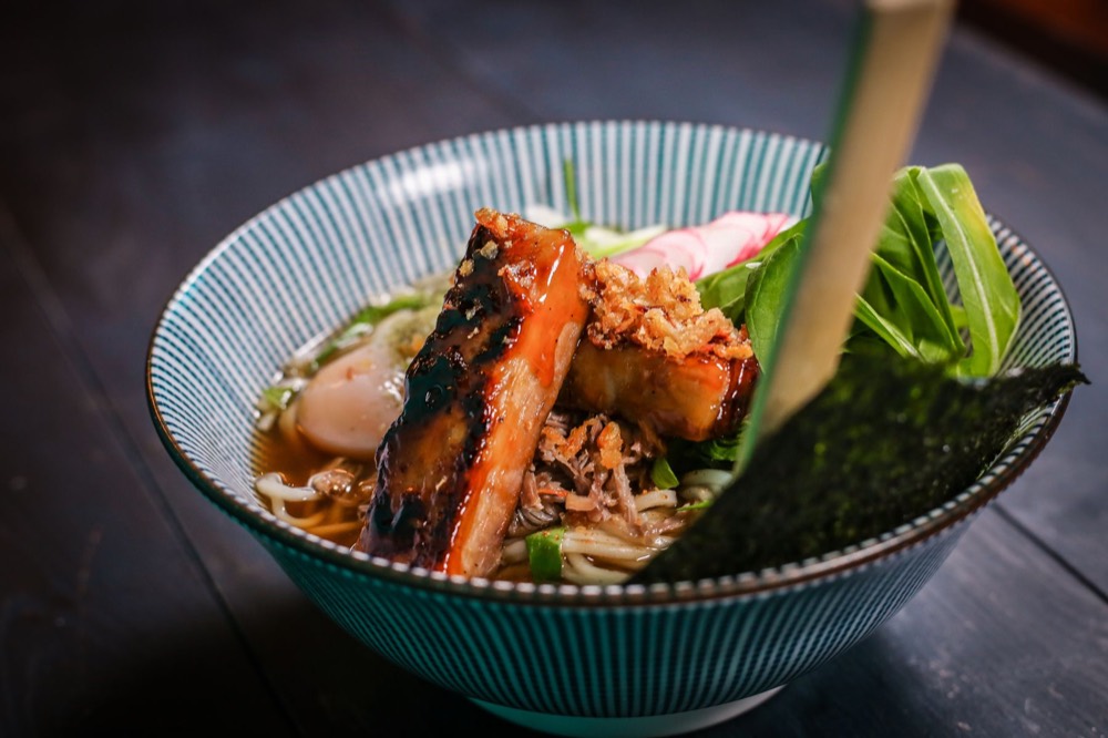 A bowl of ramen with grilled meat, greens and a soft egg, garnished with nori.
