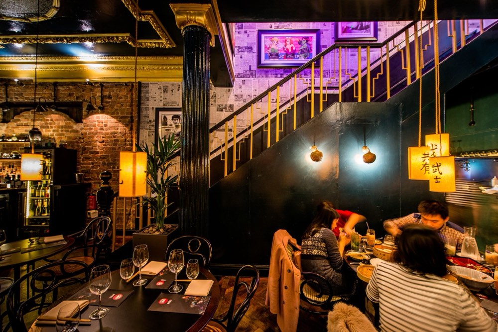 Diners at an ornate bar with exposed brick, a gold-railed staircase and newspaper-print walls.
