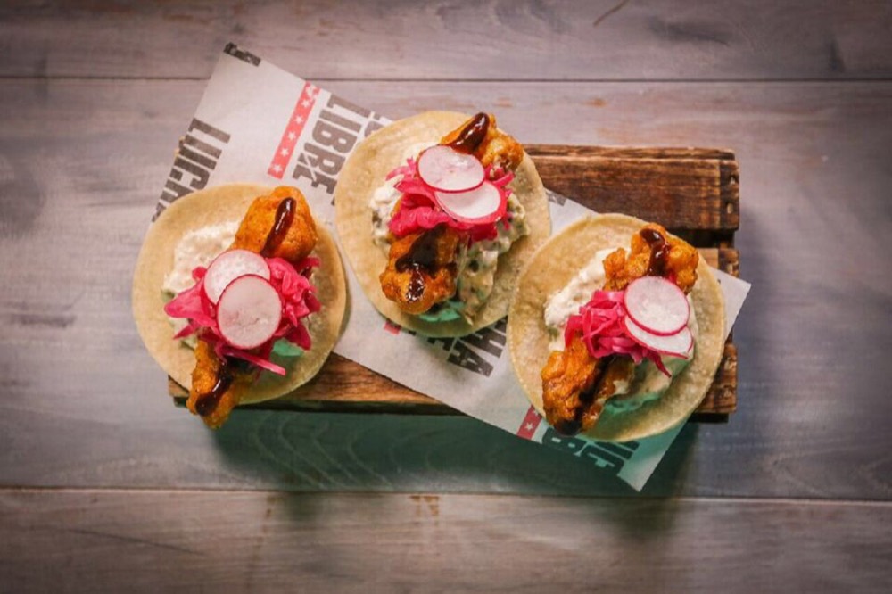 Three grilled chicken tacos topped with pink pickled onion and radish on a wooden board at Lucha Libre.