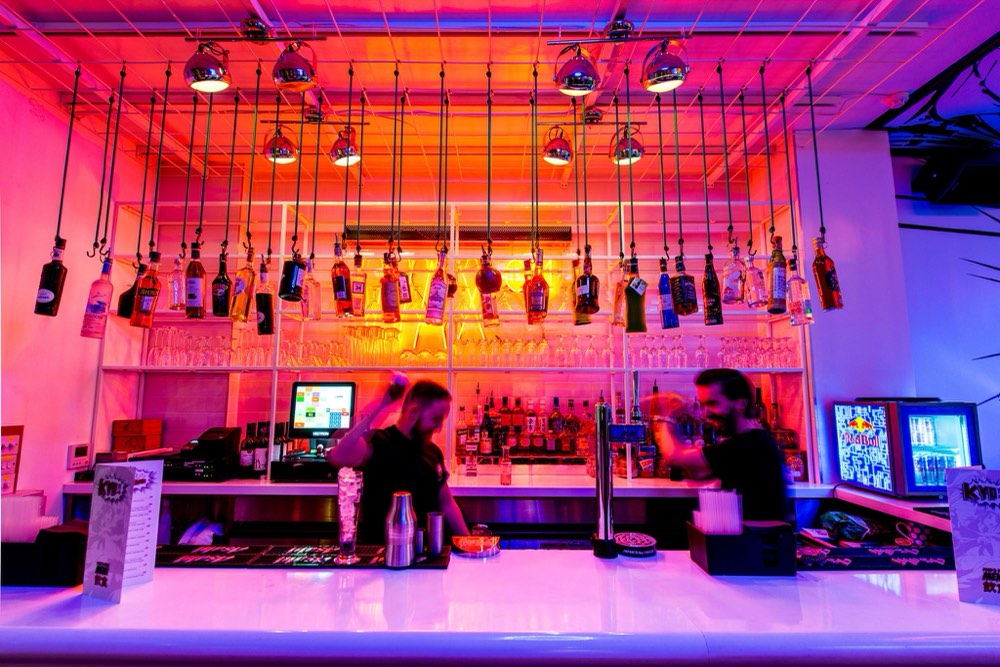 Two bartenders making drinks under a canopy of hanging spirit bottles and vivid neon lighting at a late-night bar.