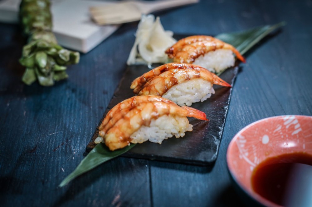 Three pieces of glazed prawn nigiri sushi on a slate board with pickled ginger and dipping sauce.