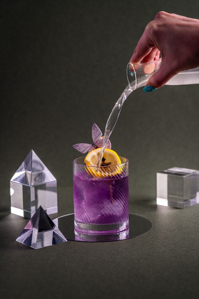 A colour-changing purple cocktail with a lemon wheel and butterfly garnish being topped from a carafe, styled with glass prisms.