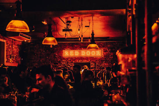 The glowing red neon Red Door sign above a busy, candle-lit bar crowd.