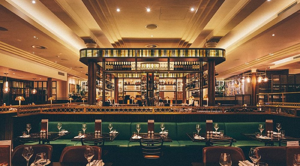 An elegant restaurant dining room with green banquette seating, laid tables and a central drinks display.