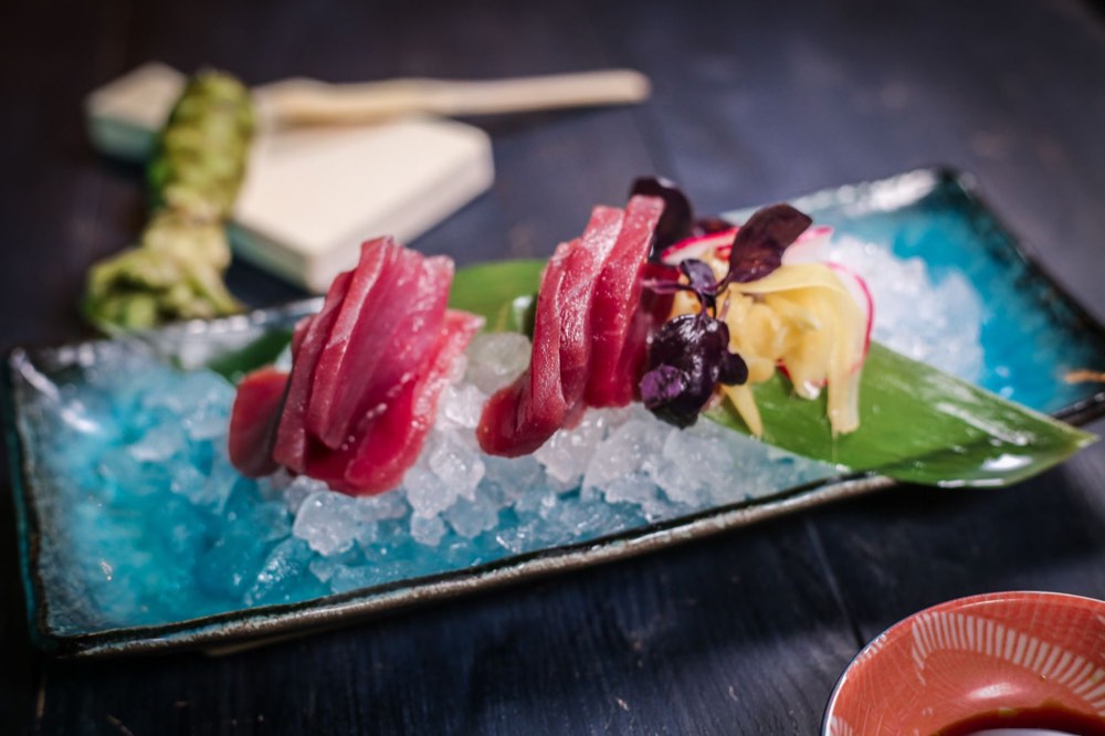 Slices of fresh tuna sashimi on crushed ice over a turquoise plate, with wasabi, pickled ginger and radish.
